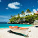 Colourful fishing boats resting on a quiet Caribbean beach with turquoise water, palm trees, and cliffside houses under a bright blue sky, reflecting a peaceful heritage travel atmosphere.