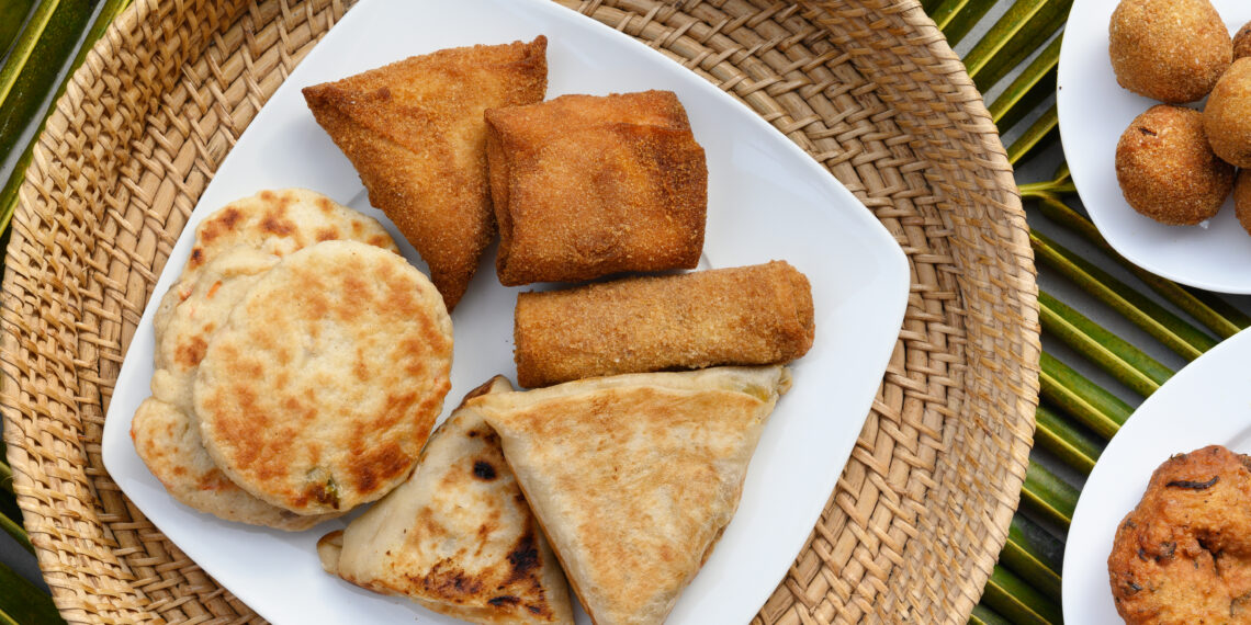 Assorted Indo-Caribbean style fried snacks and flatbreads including stuffed roti parcels and savoury pastries arranged on a white plate over a woven mat, reflecting regional street-food influences similar to Guyana and Trinidad roti traditions.