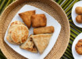 Assorted Indo-Caribbean style fried snacks and flatbreads including stuffed roti parcels and savoury pastries arranged on a white plate over a woven mat, reflecting regional street-food influences similar to Guyana and Trinidad roti traditions.