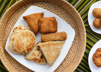 Assorted Indo-Caribbean style fried snacks and flatbreads including stuffed roti parcels and savoury pastries arranged on a white plate over a woven mat, reflecting regional street-food influences similar to Guyana and Trinidad roti traditions.