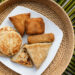 Assorted Indo-Caribbean style fried snacks and flatbreads including stuffed roti parcels and savoury pastries arranged on a white plate over a woven mat, reflecting regional street-food influences similar to Guyana and Trinidad roti traditions.
