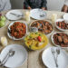 Family-style Sunday lunch table featuring a variety of Indo-Caribbean dishes including curry chicken, fried vegetables, savoury snacks, dates, and fresh fruit, with empty plates set for a shared meal.