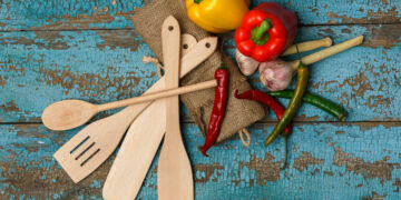 Traditional Caribbean cooking tools including a tawa, belna rolling pin, karahi pot, sil batta grinding stone, and wooden utensils arranged on a rustic kitchen surface.