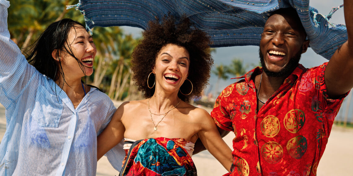 Three young adults laughing together on a sunny beach, holding a flowing scarf above their heads, capturing a joyful multicultural travel moment that reflects the vibrancy and global connection often celebrated in Indo-Caribbean culture online.