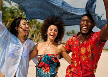Three young adults laughing together on a sunny beach, holding a flowing scarf above their heads, capturing a joyful multicultural travel moment that reflects the vibrancy and global connection often celebrated in Indo-Caribbean culture online.