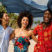 Three young adults laughing together on a sunny beach, holding a flowing scarf above their heads, capturing a joyful multicultural travel moment that reflects the vibrancy and global connection often celebrated in Indo-Caribbean culture online.