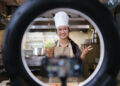 Young home cook filming a recipe video through a ring light while holding fresh vegetables in a modern kitchen.