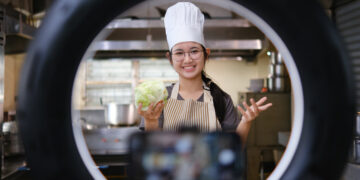 Young home cook filming a recipe video through a ring light while holding fresh vegetables in a modern kitchen.