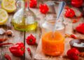 Jar of homemade Caribbean pepper sauce made with red chillies and habanero peppers on a rustic wooden table, surrounded by fresh peppers, garlic, lemon, and oil.