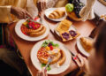A multicultural breakfast table featuring sausages, baked beans, toast with jam, croissants, fresh fruit, and salad, as two people enjoy a hearty meal together in a warm dining setting.