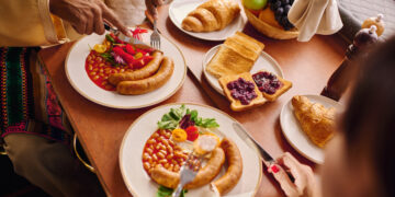 A multicultural breakfast table featuring sausages, baked beans, toast with jam, croissants, fresh fruit, and salad, as two people enjoy a hearty meal together in a warm dining setting.