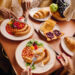 A multicultural breakfast table featuring sausages, baked beans, toast with jam, croissants, fresh fruit, and salad, as two people enjoy a hearty meal together in a warm dining setting.