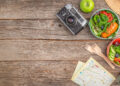 Travel-friendly healthy meal setup on a rustic wooden table with fresh salad bowls, tea, a vintage camera, reusable flask, and map for outdoor adventure inspiration.