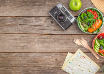Travel-friendly healthy meal setup on a rustic wooden table with fresh salad bowls, tea, a vintage camera, reusable flask, and map for outdoor adventure inspiration.