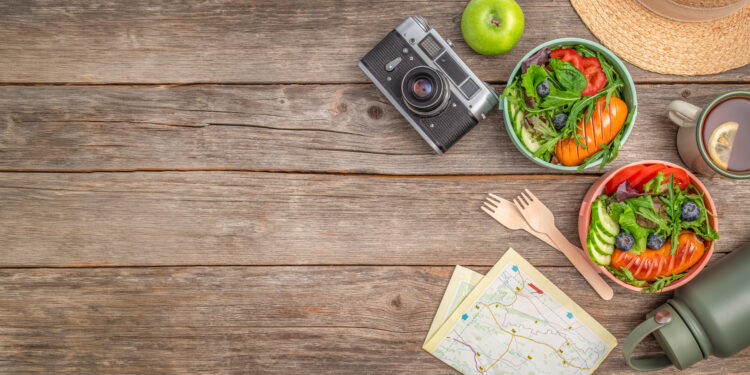 Travel-friendly healthy meal setup on a rustic wooden table with fresh salad bowls, tea, a vintage camera, reusable flask, and map for outdoor adventure inspiration.