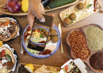 Overhead view of Mediterranean-style vegetarian street food with falafel wraps, spices, fresh vegetables, and a person photographing the meal on a smartphone.