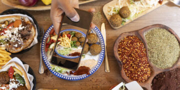 Overhead view of Mediterranean-style vegetarian street food with falafel wraps, spices, fresh vegetables, and a person photographing the meal on a smartphone.