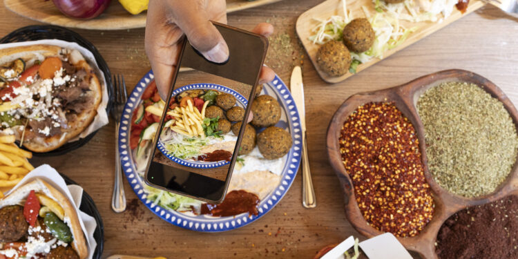 Overhead view of Mediterranean-style vegetarian street food with falafel wraps, spices, fresh vegetables, and a person photographing the meal on a smartphone.