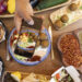 Overhead view of Mediterranean-style vegetarian street food with falafel wraps, spices, fresh vegetables, and a person photographing the meal on a smartphone.