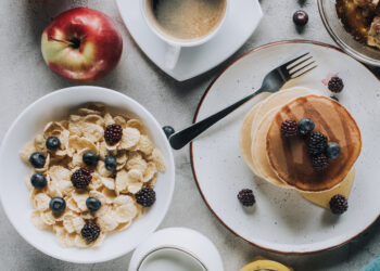 Top-view breakfast spread featuring pancakes with berries, cereal, coffee, milk, apples, honeycomb, and orange juice on a light table.