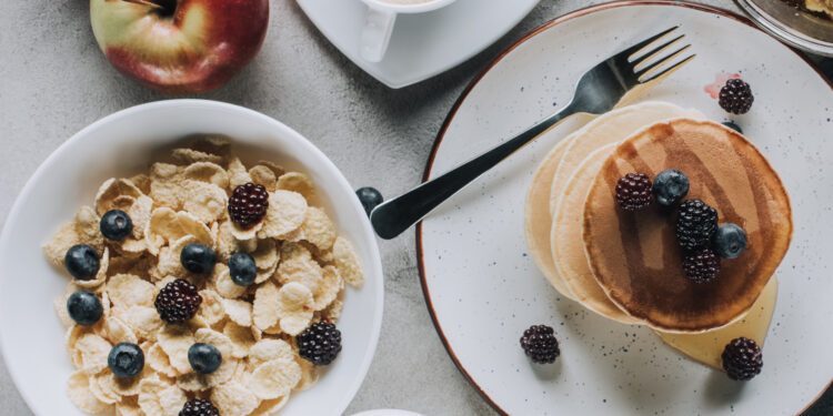 Top-view breakfast spread featuring pancakes with berries, cereal, coffee, milk, apples, honeycomb, and orange juice on a light table.