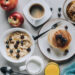Top-view breakfast spread featuring pancakes with berries, cereal, coffee, milk, apples, honeycomb, and orange juice on a light table.