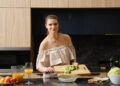 A woman preparing a fresh homemade salad in a modern kitchen, chopping cucumbers on a wooden board beside bowls of vegetables and a glass of orange juice.