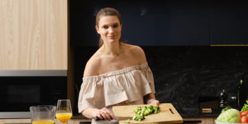 A woman preparing a fresh homemade salad in a modern kitchen, chopping cucumbers on a wooden board beside bowls of vegetables and a glass of orange juice.